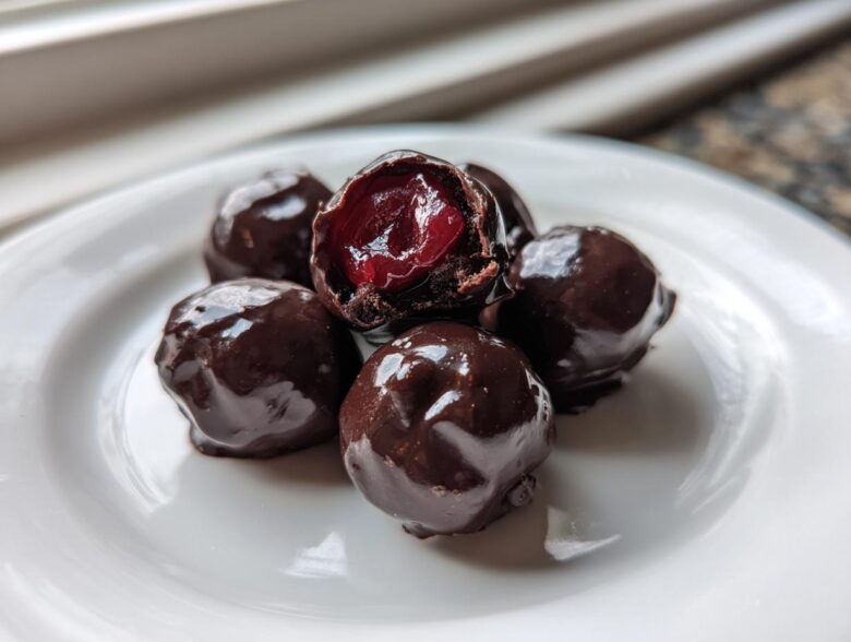Close-up of five homemade chocolate covered cherries on a white plate, one is broken open showing the red cherry inside.
