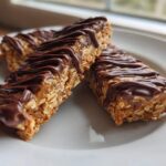 Three homemade protein bars made with oats and drizzled generously with melted dark chocolate, served on a white plate.