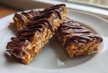 Three homemade protein bars made with oats and drizzled generously with melted dark chocolate, served on a white plate.
