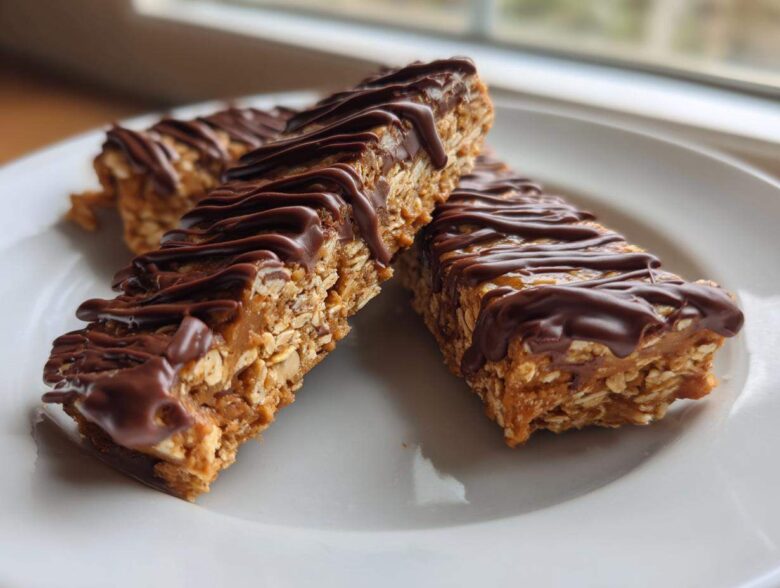 Three homemade protein bars made with oats and drizzled generously with melted dark chocolate, served on a white plate.