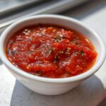 A small white bowl filled with rich, chunky homemade pizza sauce seasoned with herbs and spices.