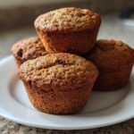 A close-up of four freshly baked honey bran muffins stacked slightly on a white plate.