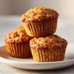 Close-up of three perfectly baked honey bran muffins stacked on a rustic white plate.