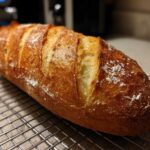 A golden-brown, freshly baked Italian bread loaf with deep scores resting on a wire cooling rack.