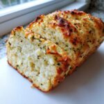 A freshly baked loaf of Italian herb and cheese bread, partially sliced, showing melted cheese pockets and green herbs.