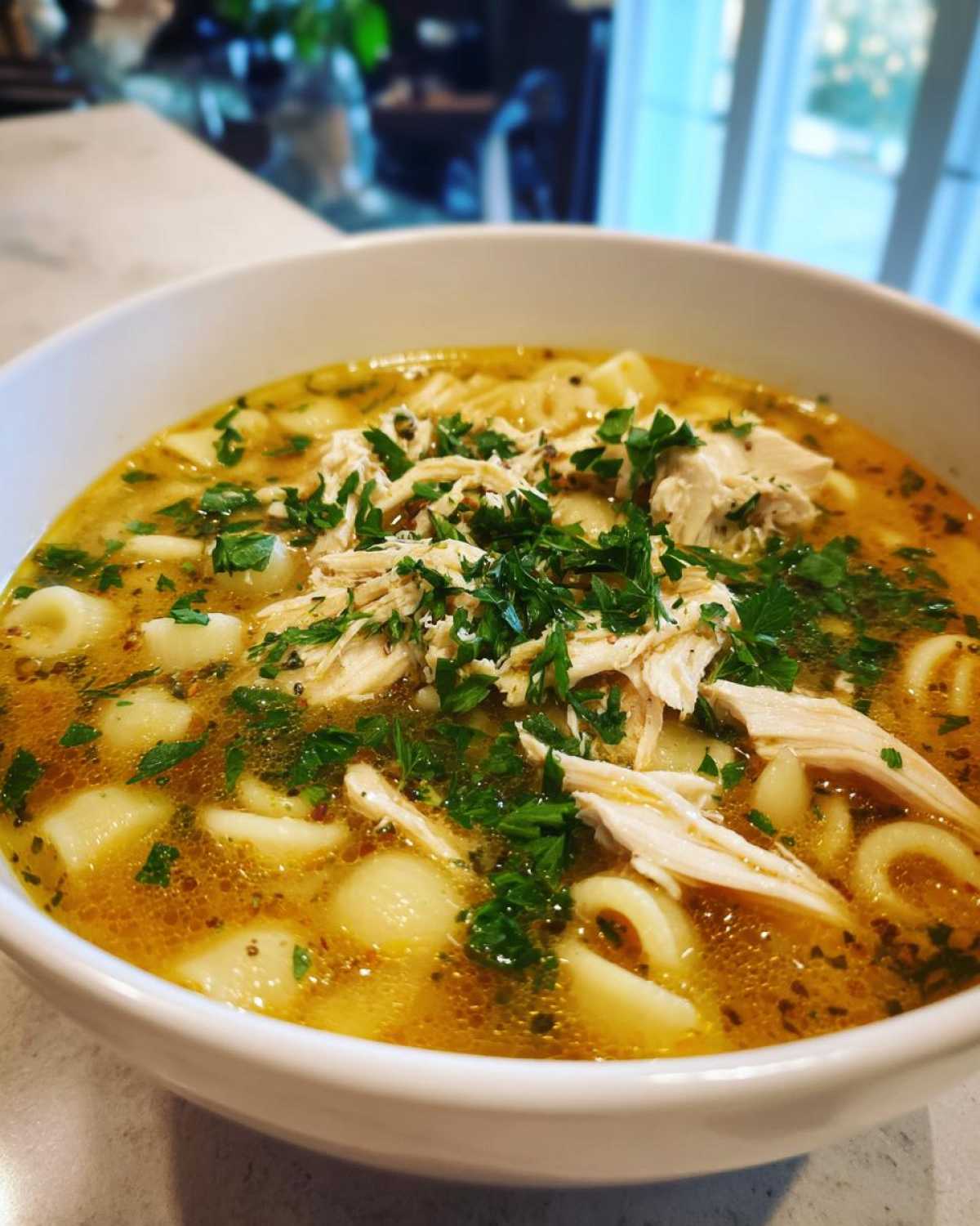 Close-up of a white bowl filled with rich, golden Italian penicillin soup, featuring shredded chicken and pasta, topped with fresh parsley.
