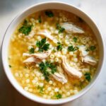 A close-up overhead view of a white bowl filled with steaming italian penicillin soup, featuring shredded chicken, small pasta, and fresh parsley.