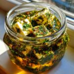 Close-up of white cheese cubes soaking in oil and herbs for marinated cheese in a clear glass jar.
