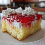 Close-up of a moist slice of jello poke cake featuring a yellow sponge base, white filling, bright red gelatin topping, and dollops of whipped cream.