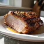 Close-up of a perfectly cooked, juicy pork loin roast slice with a dark, herb-crusted exterior resting in its own juices.