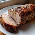 A perfectly cooked, juicy pork loin roast with a browned, herb-crusted exterior, resting on a white plate with juices.