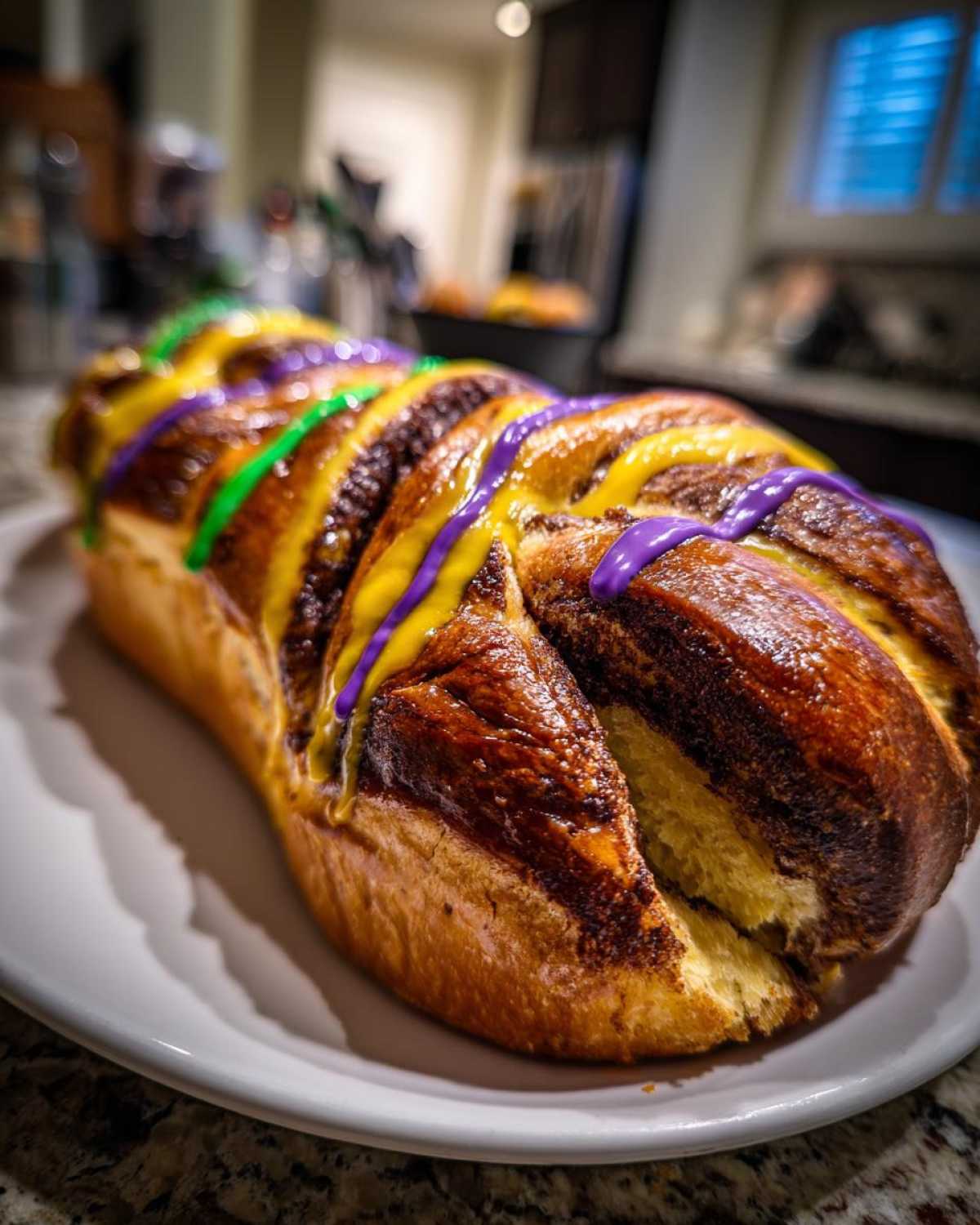 A freshly baked, braided king cake loaf drizzled with purple, yellow, and green icing, resting on a white plate.
