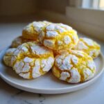 A stack of vibrant yellow lemon crinkle cookies heavily coated in white powdered sugar, resting on a white plate.