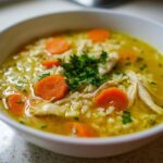 Close-up of a white bowl filled with Lemon Ginger Turmeric Chicken and Rice Soup, featuring shredded chicken, rice, sliced carrots, and parsley.