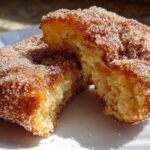 A close-up of a Mexican buñuelos pastry, broken in half to show the fluffy interior, coated heavily in cinnamon sugar.