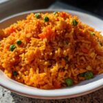 A close-up of a mound of vibrant orange Mexican rice recipe served in a white bowl, garnished with green peas.