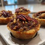Close-up of freshly baked mini pecan pies with glossy filling and whole pecans, resting in a mini muffin tin.