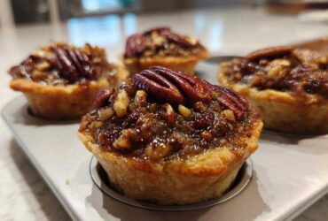 Close-up of freshly baked mini pecan pies with glossy filling and whole pecans, resting in a mini muffin tin.