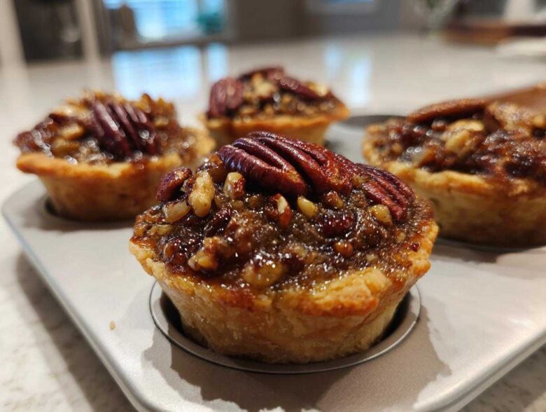 Close-up of freshly baked mini pecan pies with glossy filling and whole pecans, resting in a mini muffin tin.
