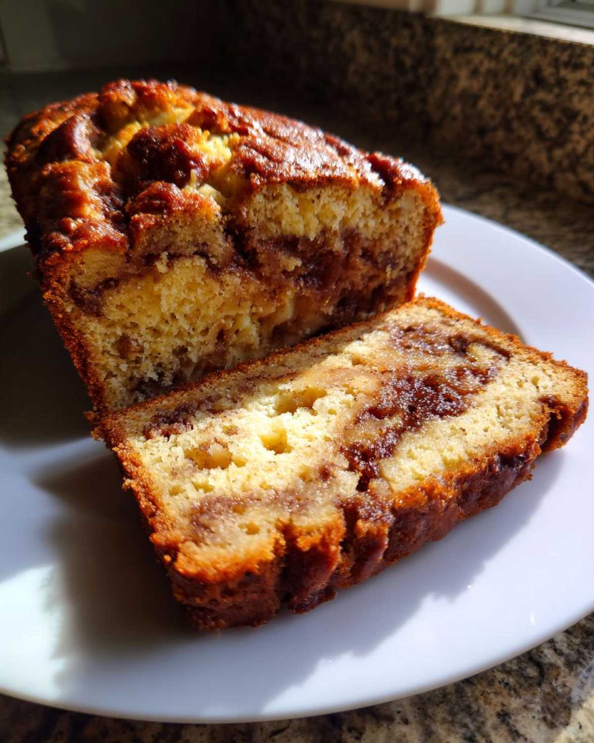 A close-up of moist apple bread with a rich cinnamon swirl, sliced on a white plate.