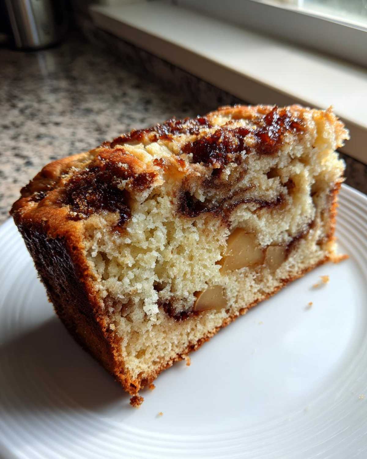 A close-up slice of moist apple bread showing chunks of apple and a dark cinnamon swirl topping.