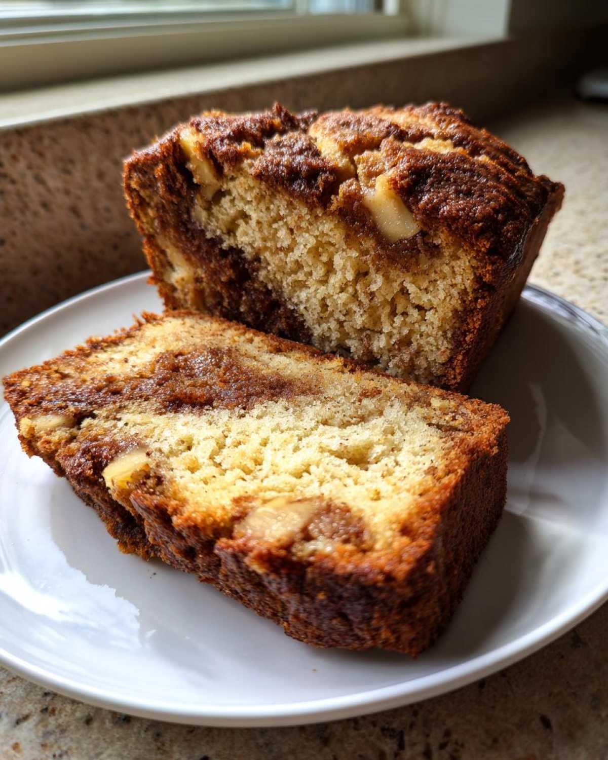 Close-up of moist apple bread loaf sliced on a white plate, showing cinnamon swirl and apple chunks.