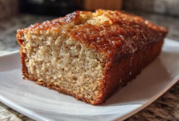 Close-up of a slice of moist banana bread with a shiny, caramelized glaze on top, served on a white plate.