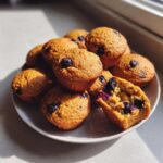 A stack of moist blueberry protein muffins on a plate, with one muffin broken open showing the soft interior.
