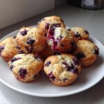 A pile of golden-brown blueberry protein muffins, one broken open to show the moist interior, served on a white plate.