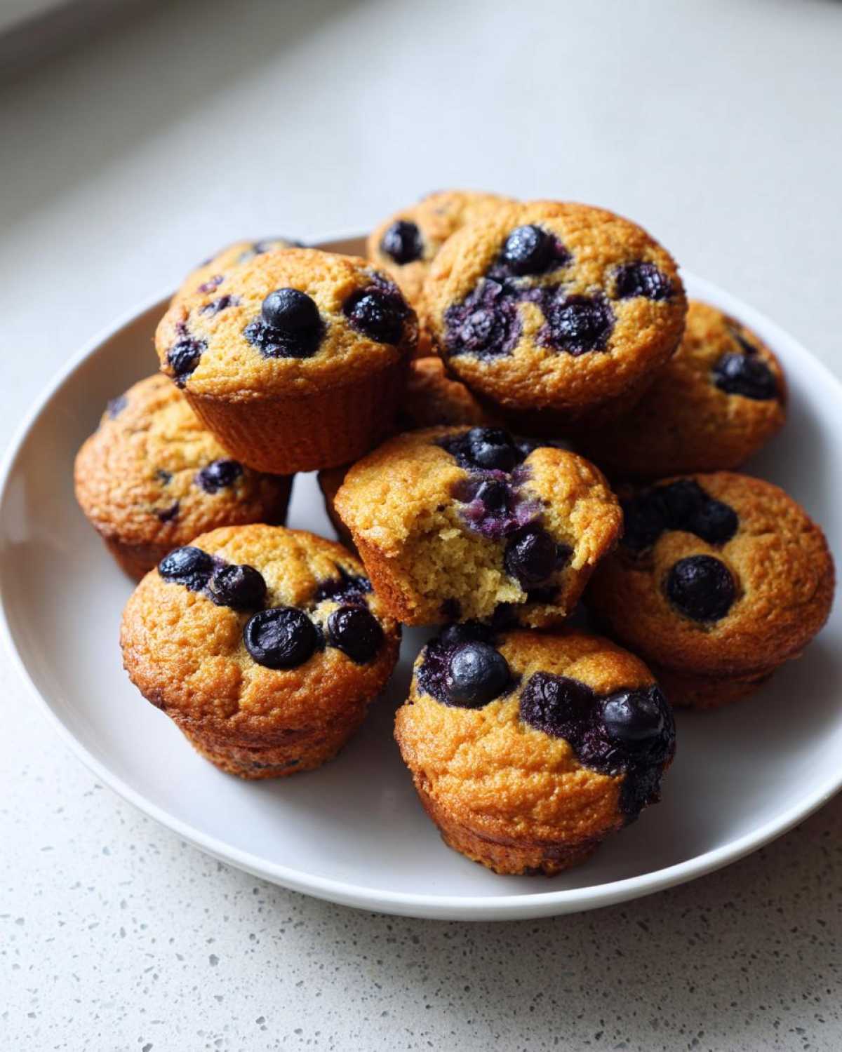 A pile of golden brown blueberry protein muffins stacked on a white plate, one muffin has a bite taken out.