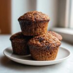 A stack of three dark, moist bran muffins with textured tops sitting on a small white plate.