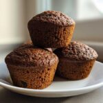 Three dark brown, moist bran muffins stacked on a small white plate, backlit by a window.