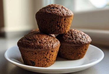 Three dark brown, moist bran muffins stacked on a small white plate, backlit by a window.