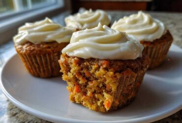 Close-up of four carrot cake muffins with thick cream cheese frosting, one muffin is broken open showing shredded carrots.