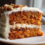 Close-up of a moist, two-layer carrot cake recipe slice with thick cream cheese frosting and chopped walnuts.