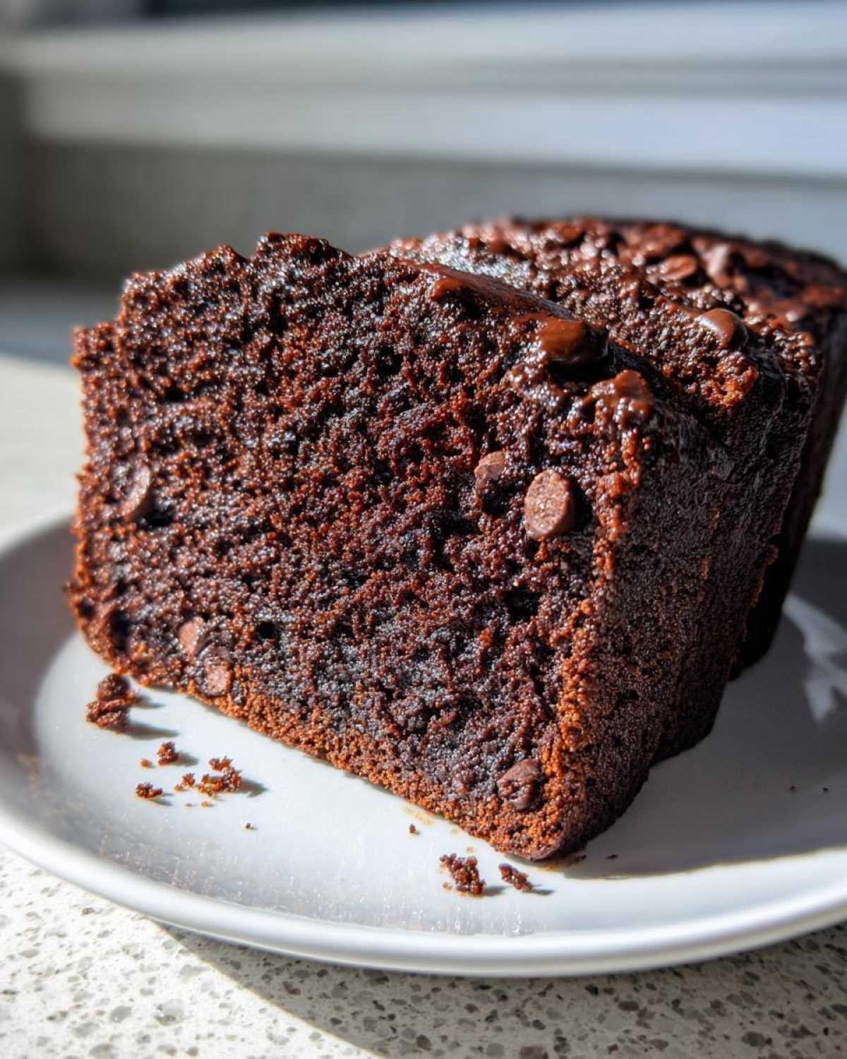 Close-up of a thick, moist slice of chocolate banana bread studded with chocolate chips, sitting on a white plate.