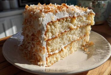 A generous slice of moist coconut cake with white frosting and toasted coconut flakes on a cream plate.