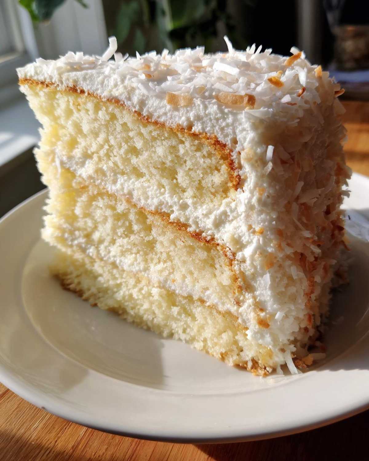 A tall slice of moist coconut cake with white frosting and toasted coconut flakes on top.