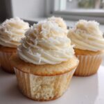 Close-up of several moist coconut cupcakes topped with white frosting and shredded coconut.