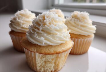 Close-up of several moist coconut cupcakes topped with white frosting and shredded coconut.