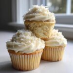 Three moist coconut cupcakes stacked, topped with thick white frosting and shredded coconut flakes.
