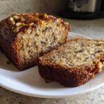 A freshly baked date nut bread loaf, partially sliced, topped with walnuts, sitting on a white plate.