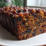 Close-up of a thick, moist slice of dark fruit cake loaded with dried fruits and nuts on a white plate.