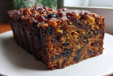 Close-up of a thick, moist slice of dark fruit cake loaded with dried fruits and nuts on a white plate.