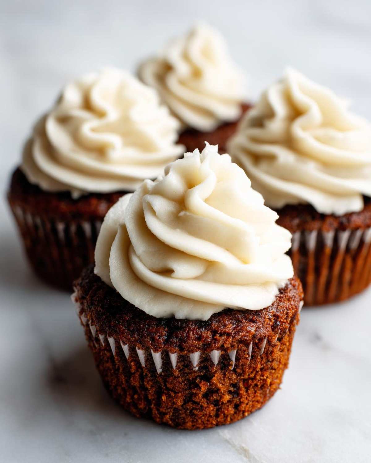 Close-up of moist gingerbread cupcakes topped with swirls of creamy white frosting on a marble surface.