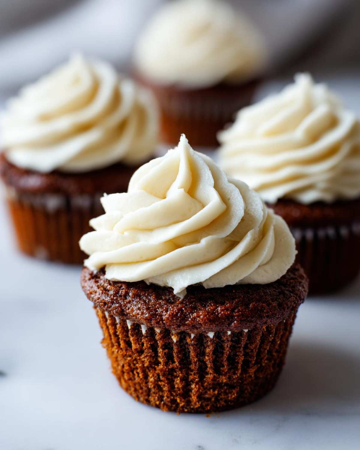 A close-up of a perfectly frosted gingerbread cupcake with creamy white frosting, with more gingerbread cupcakes blurred in the background.