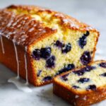 Close-up of moist blueberry bread loaf drizzled with white lemon glaze, showing juicy blueberries inside.