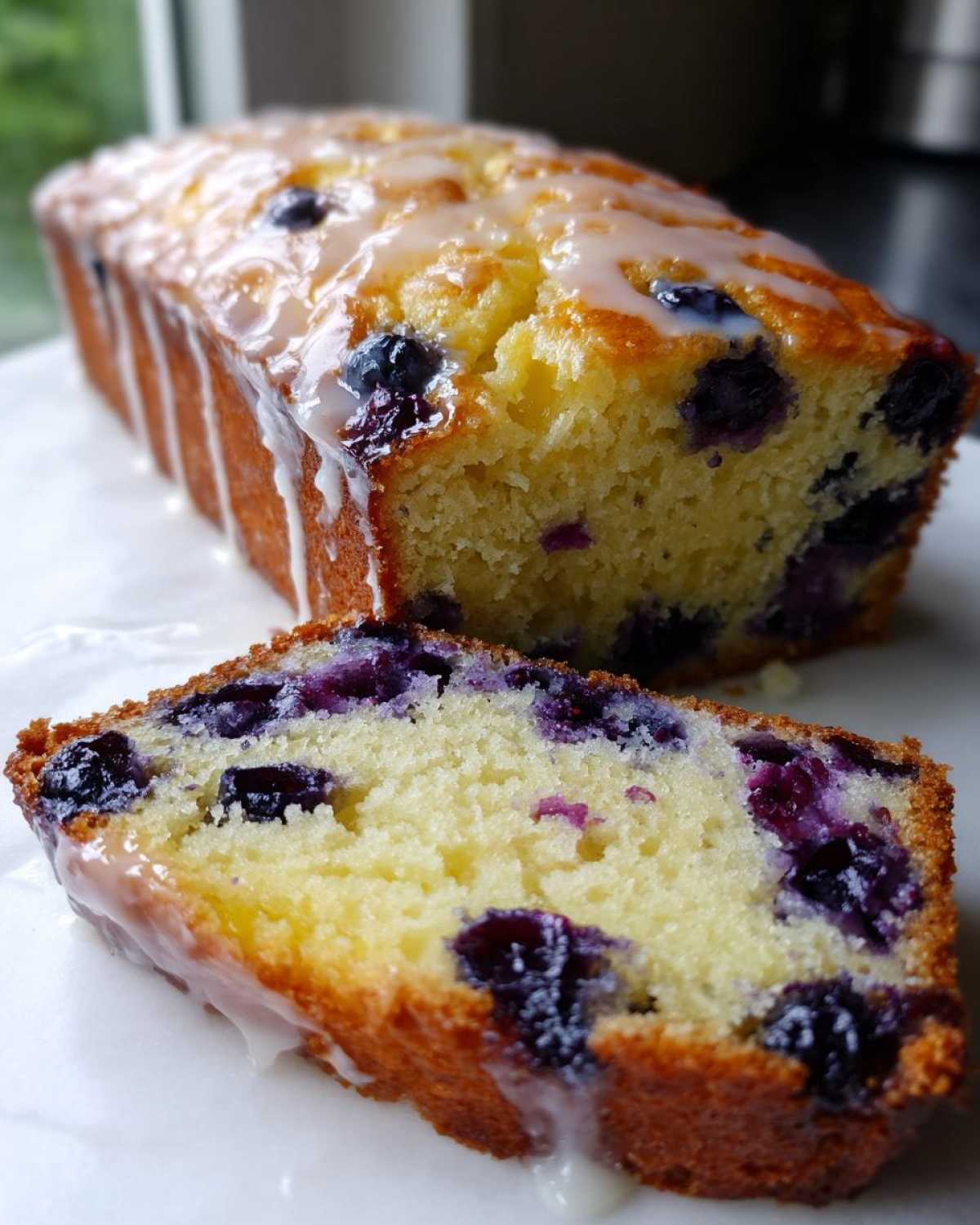 A close-up of a slice of moist lemon blueberry bread with a sweet glaze drizzled over the top.