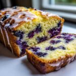 A close-up of moist lemon blueberry bread, sliced to show the interior packed with blueberries and topped with a white glaze.