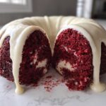 A moist red velvet bundt cake, cut in half to show the interior texture and cream cheese swirl, topped with dripping white glaze.